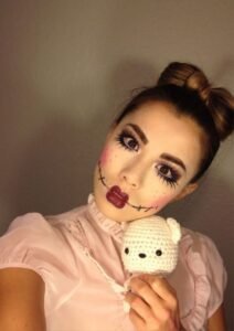 Woman wearing creepy doll halloween makeup design with bright blush, red lips, and cracked-cheek details.