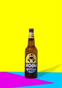 A bottle of Bira 91 BOOM Super Strong beer with a monkey mascot on the label, set against a vivid yellow, blue, and pink graphic background, highlighting the innovative design of Indian Beer Brands.