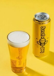 A yellow Bee Young Crafted Strong Beer can and a glass of foamy, light-colored beer on a bright yellow surface, showcasing the vibrant packaging of modern Indian Beer Brands.