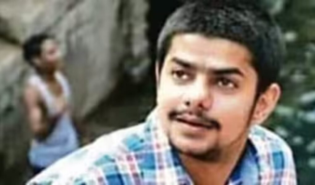 Close-up headshot of a young man with a mustache wearing a plaid shirt, identified as an associate or image related to Anmol Bishnoi in the ongoing gang investigation.