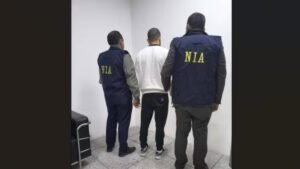 Two NIA officers, wearing official navy vests with "NIA" in yellow letters on the back, standing with a suspect believed to be **Anmol Bishnoi** after his arrest following deportation from the US.