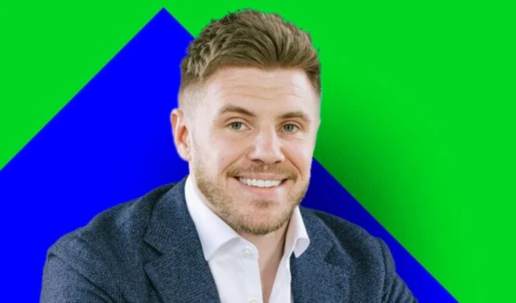 A smiling, light-skinned man with short brown hair and a stubble beard, wearing a dark blue blazer over a white shirt, set against a geometric background of bright green and blue. The background colors are bold and non-standard for a business portrait. This individual is associated with Wingstop.