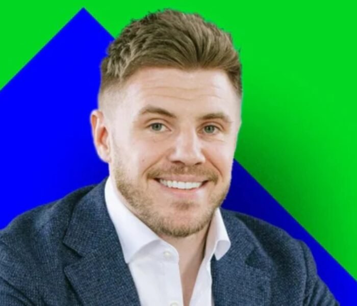 A smiling, light-skinned man with short brown hair and a stubble beard, wearing a dark blue blazer over a white shirt, set against a geometric background of bright green and blue. The background colors are bold and non-standard for a business portrait. This individual is associated with Wingstop.