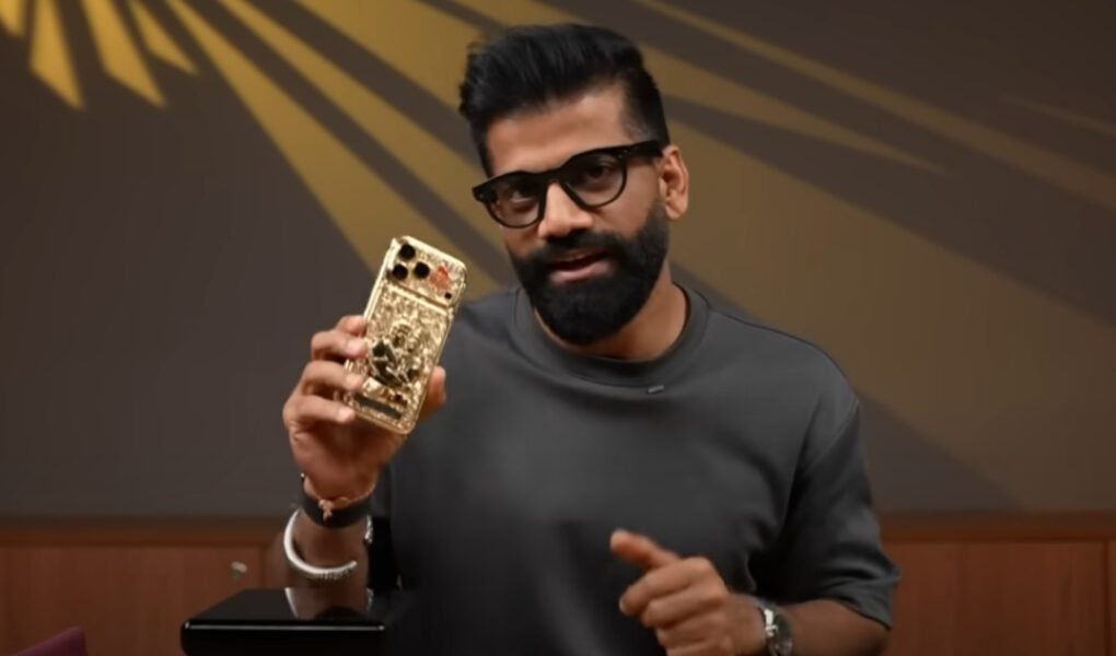 A man with a beard and glasses holding up a custom-made, gold-plated iPhone 17 Pro Max with 'Jai Shri Ram' and Ram-Hanuman carvings, which is expected to go **viral** due to its ₹50 lakh price tag.