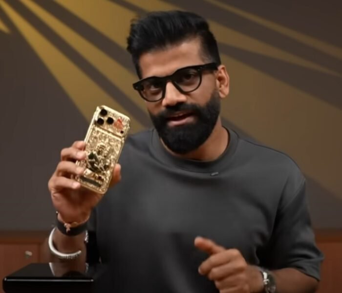 A man with a beard and glasses holding up a custom-made, gold-plated iPhone 17 Pro Max with 'Jai Shri Ram' and Ram-Hanuman carvings, which is expected to go **viral** due to its ₹50 lakh price tag.