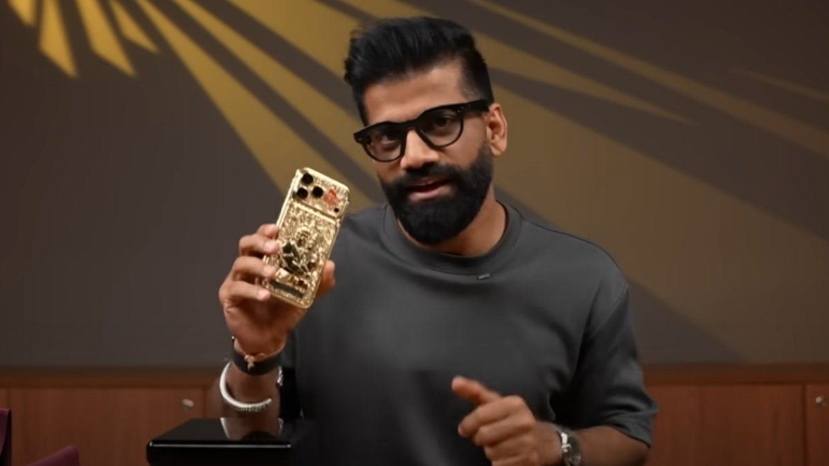 A man with a beard and glasses holding up a custom-made, gold-plated iPhone 17 Pro Max with 'Jai Shri Ram' and Ram-Hanuman carvings, which is expected to go **viral** due to its ₹50 lakh price tag.