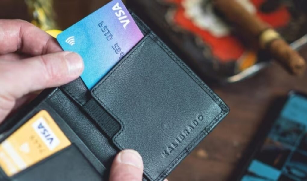 A hand taking a gradient blue and purple **Visa** card out of a black leather wallet, representing payment and travel documents in the context of the Iran entry suspension news.