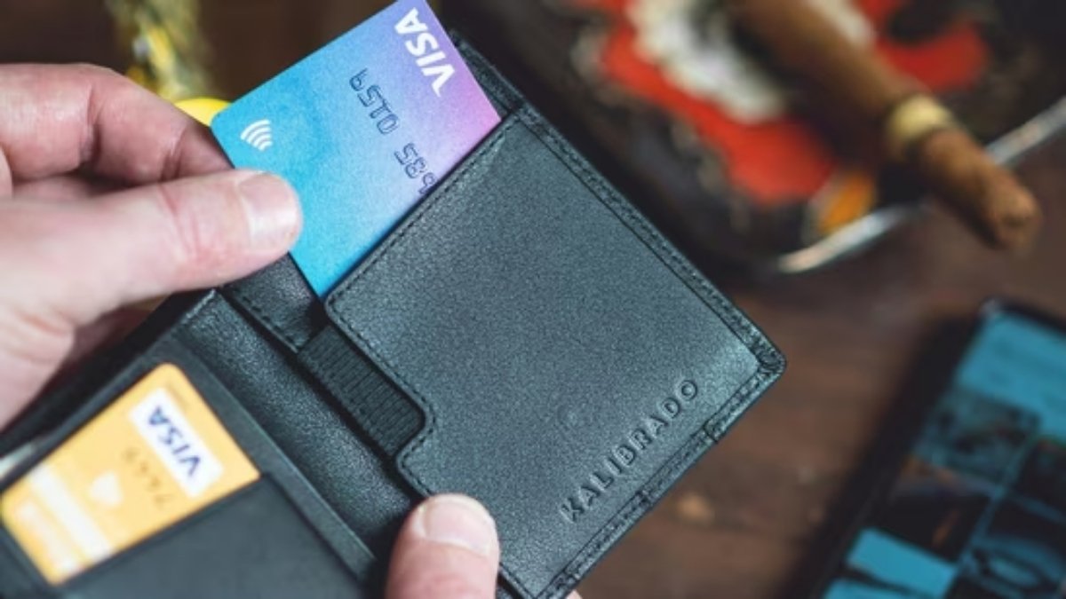 A hand taking a gradient blue and purple **Visa** card out of a black leather wallet, representing payment and travel documents in the context of the Iran entry suspension news.