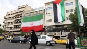 A large **Iran**ian national flag draped on a building in a city street, with people and cars passing below, symbolizing the country's travel policy change for Indian citizens.